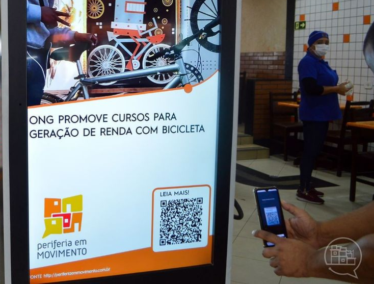 fotografia mostra pessoa apontando celular para fazer leitura de QR code em painel digital